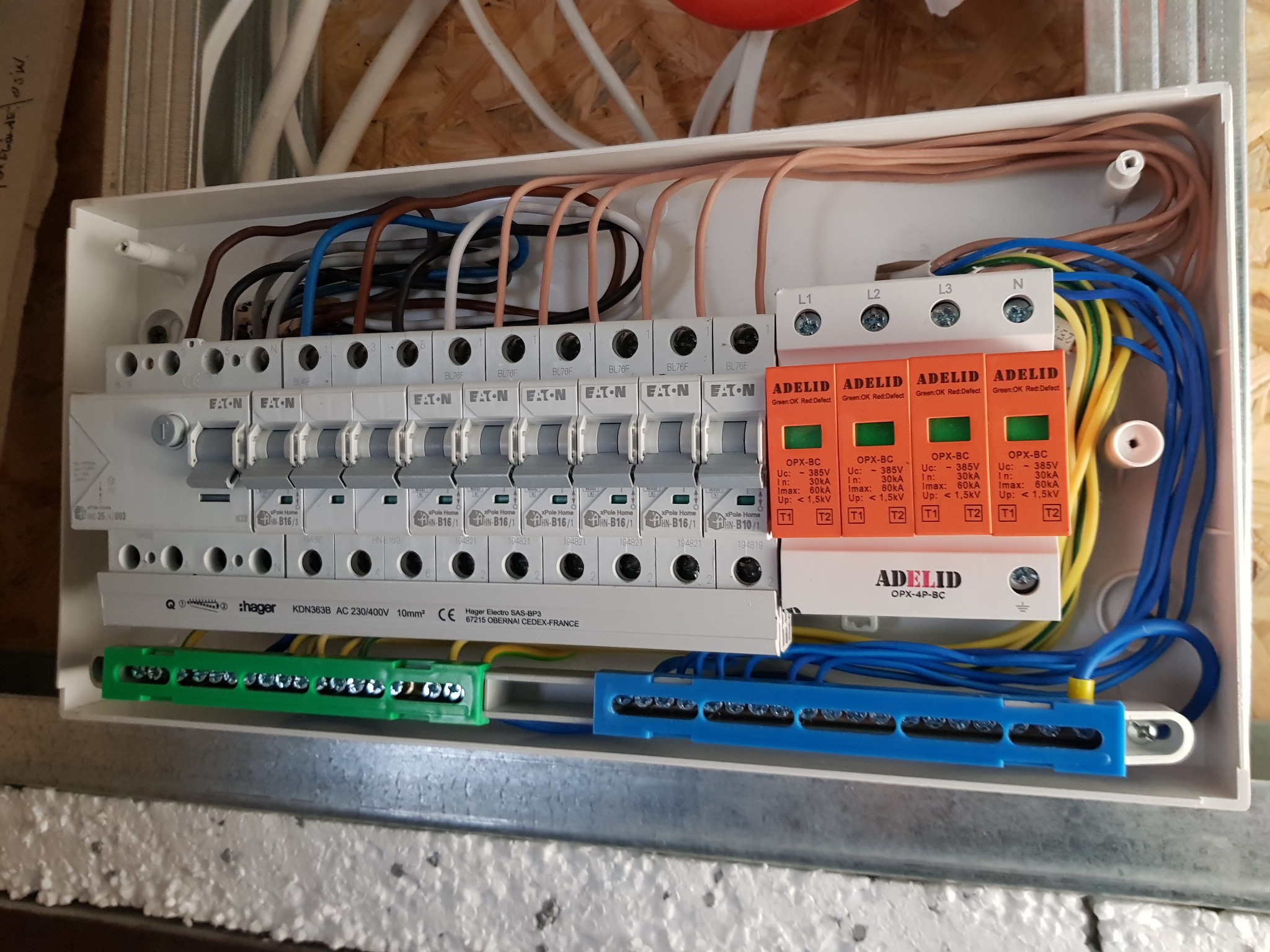 Rozdzielnica elektryczna z zabezpieczeniami przepięciowymi Adelid, widoczne bezpieczniki Eaton, przewody w kolorach niebieskim, żółtym, brązowym i białym, zamontowana w skrzynce na drewnianym podłożu.