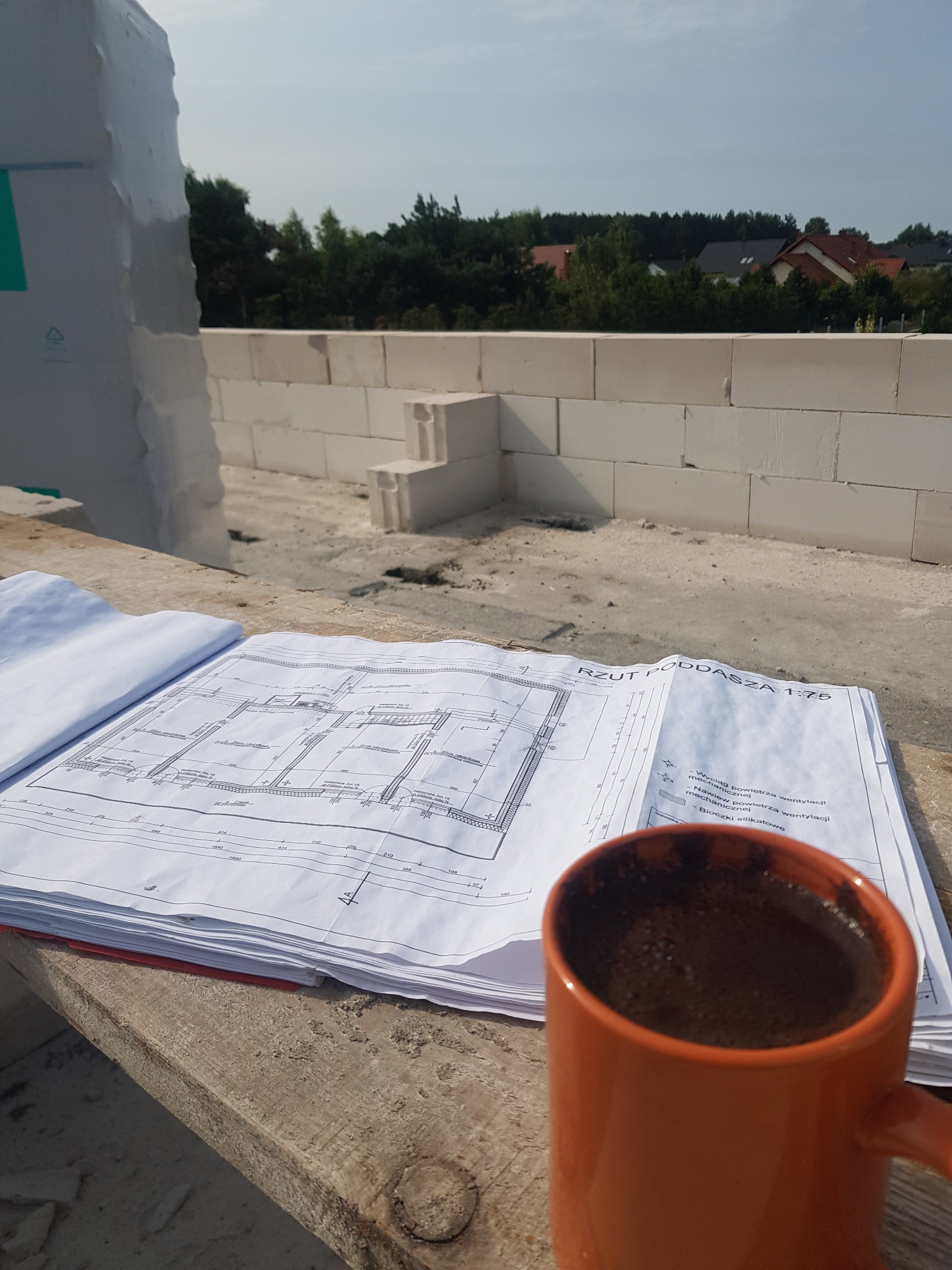 Plan rzutu poddasza 1:75 leżący na drewnianej desce obok kubka z kawą na tle budowy domu z białych bloczków silikatowych