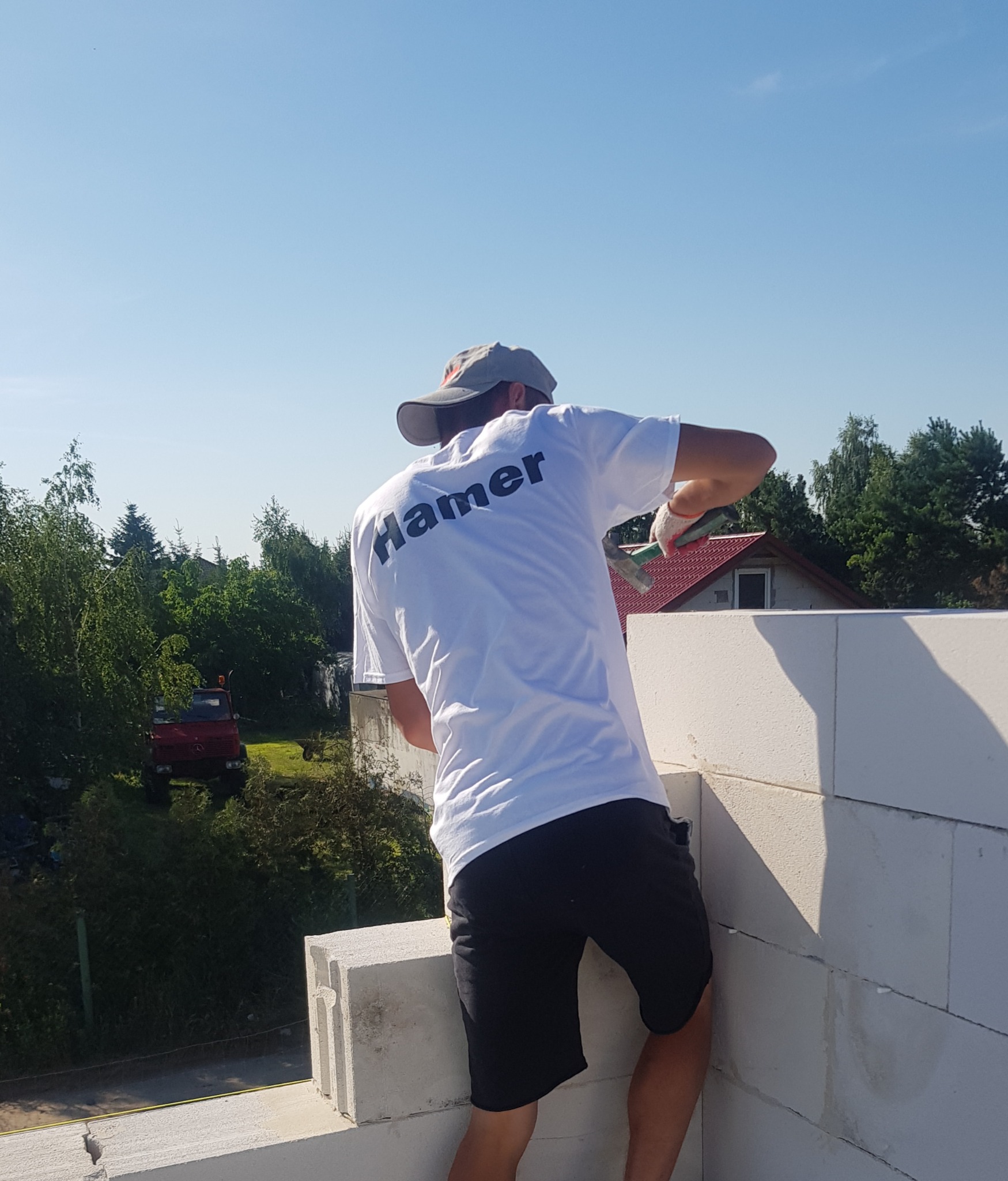 Pracownik w koszulce z logo firmy buduje ścianę z białych bloczków, w tle widoczny dom i czerwony samochód.