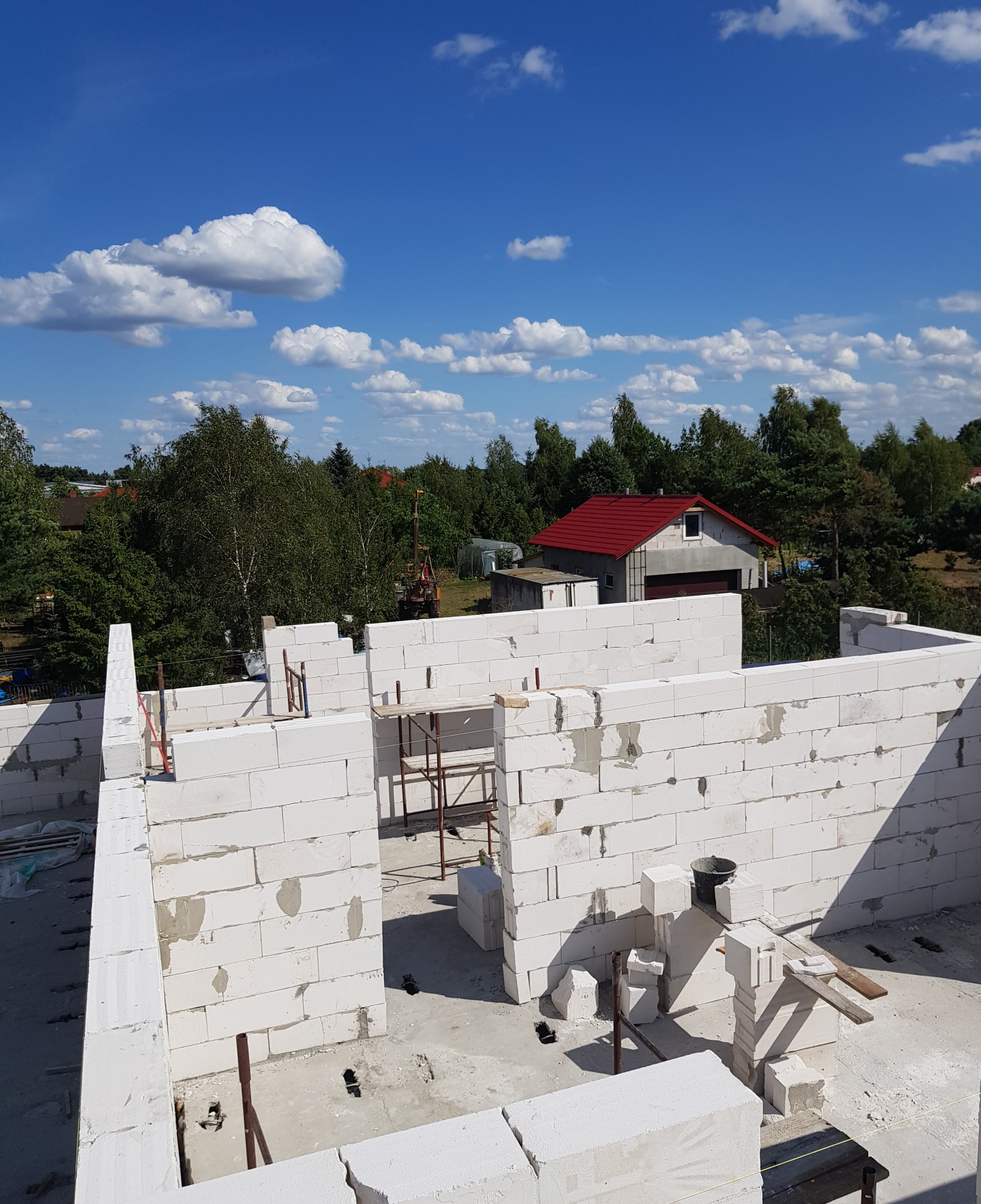 Wznoszone mury parterowego domu z białych bloczków, widoczne szalunki, rusztowania i elementy konstrukcyjne na tle błękitnego nieba z białymi chmurami oraz zabudowań gospodarczych z czerwonym dachem.
