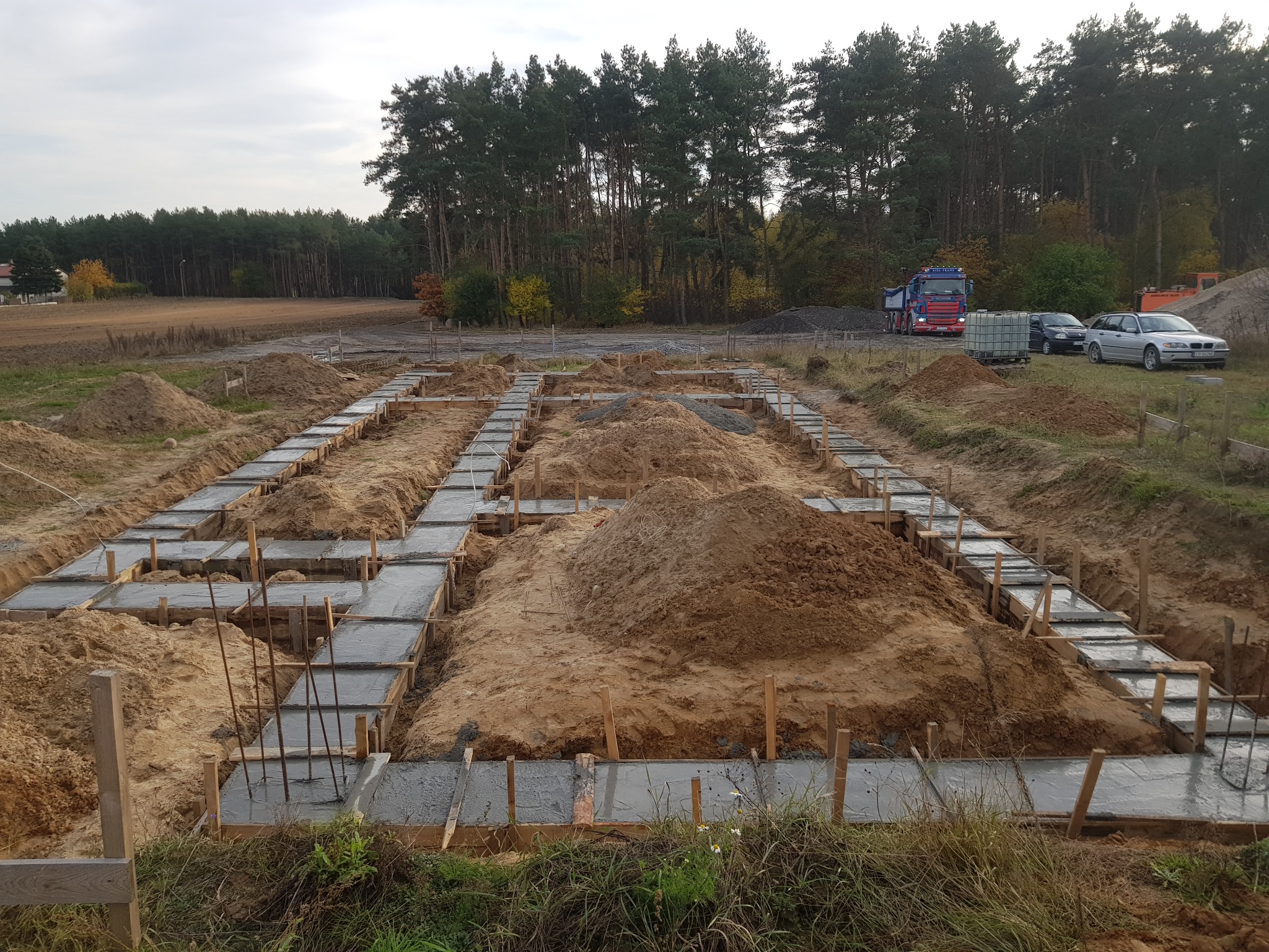 Wylane fundamenty domu murowanego z widocznym zbrojeniem, drewnianym szalunkiem, stertami piasku, ciężarówką i samochodami w tle.