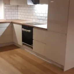 Jasna kuchnia z białymi, błyszczącymi szafkami, drewnianym blatem i białymi płytkami ułożonymi w cegiełkę; piekarnik z zabezpieczeniami na drzwiczkach.