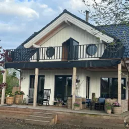 Biały dom z drewnianą elewacją, czarnym dachem i balustradą balkonową z okrągłymi oknami, widok z drewnianego tarasu z meblami ogrodowymi i kwiatami w donicach.