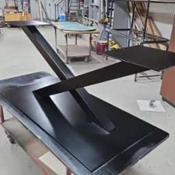 Czarny, metalowy stół z geometryczną podstawą na wózku w warsztacie, widoczne ślady obróbki.