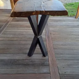 Stół z blatem z litego drewna o nieregularnym kształcie, wsparty na metalowej podstawie w kształcie litery X, ustawiony na drewnianym tarasie.