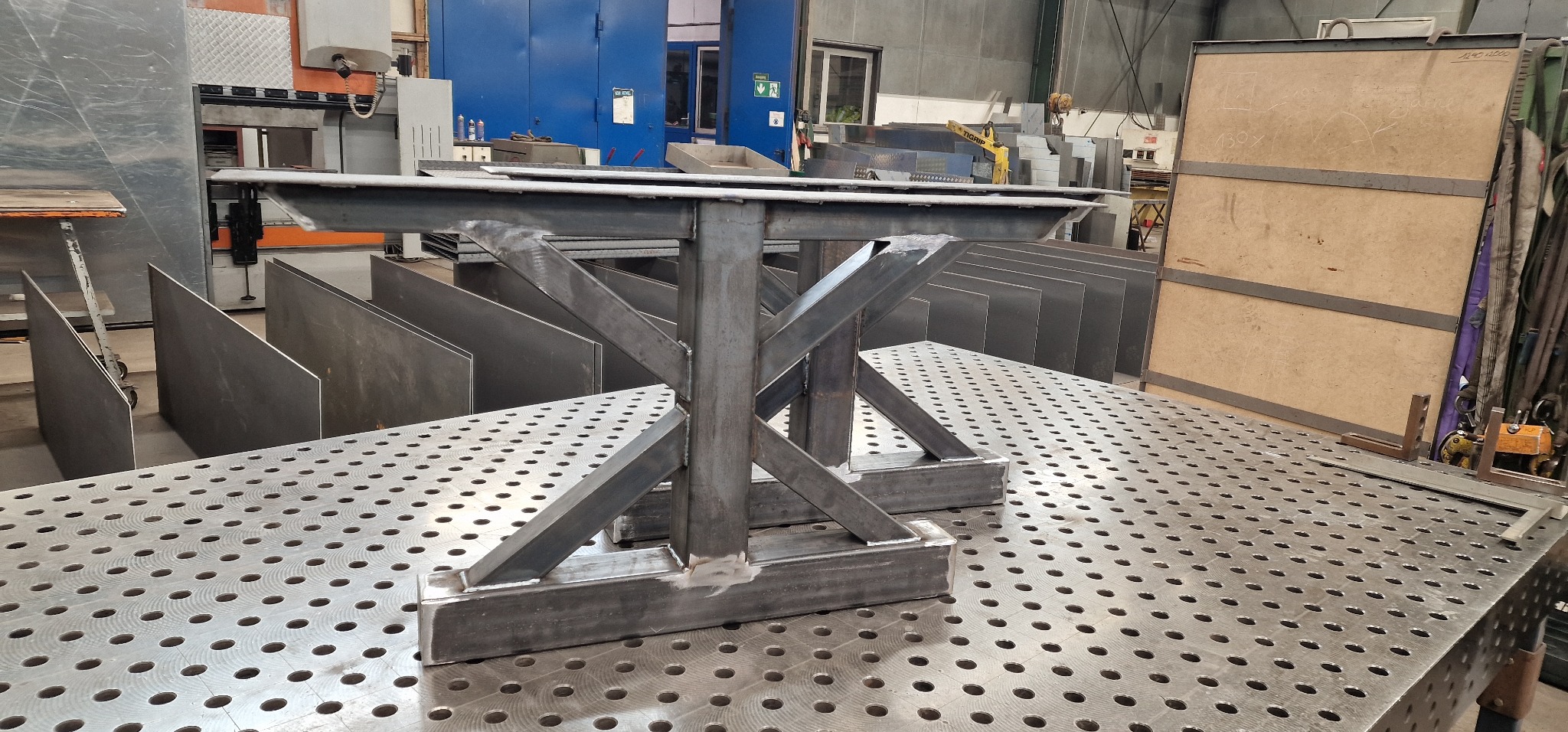 Stalowa konstrukcja stołu spawalniczego z nogami w kształcie litery X, widoczne spawy, tło warsztatowe z blachami i maszynami.