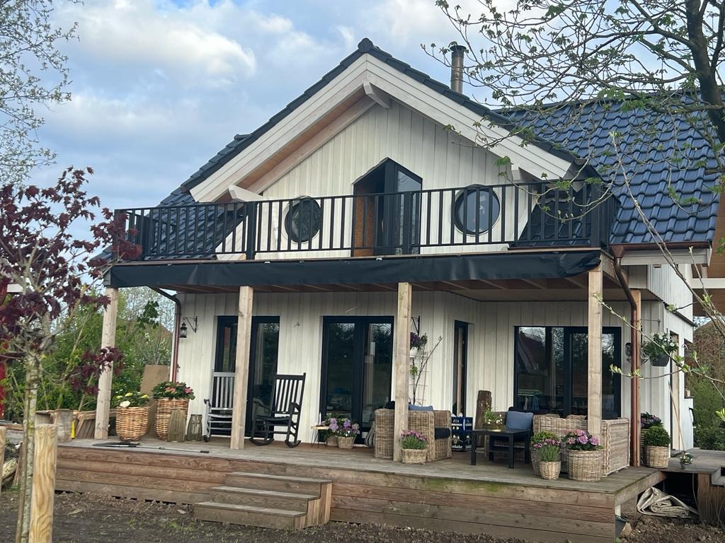 Biały dom z drewnianą elewacją, czarnym dachem i balustradą balkonową z okrągłymi oknami, widok z drewnianego tarasu z meblami ogrodowymi i kwiatami w donicach.
