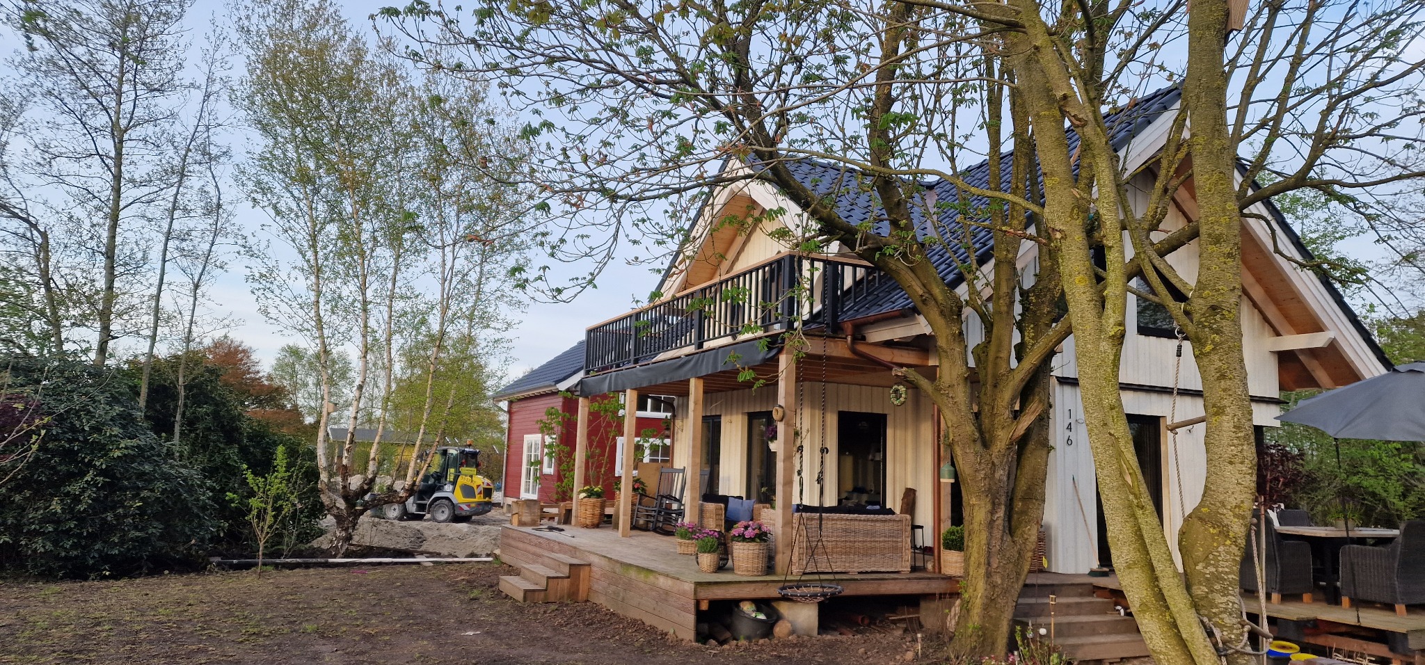 Widok domu z drewnianym tarasem i czarną balustradą balkonu, z wykopem i małą koparką w tle, sugerujący prace ziemne przed założeniem ogrodu lub montażem ogrodzenia.