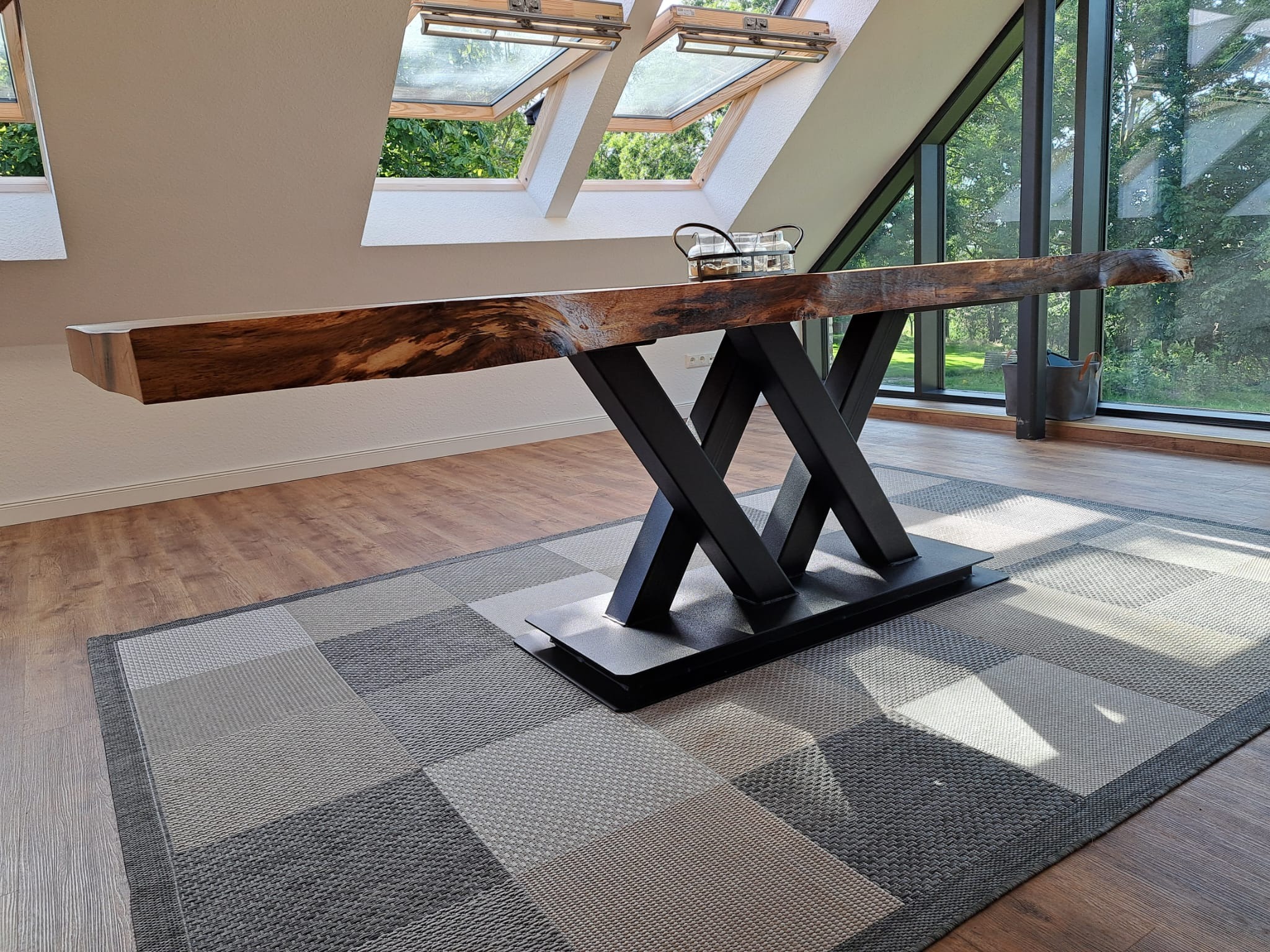 Stół z blatem z litego drewna o nieregularnych krawędziach, wsparty na czarnej, metalowej podstawie w kształcie litery X, na tle jasnego wnętrza z oknami dachowymi i dużym oknem z widokiem na zieleń.
