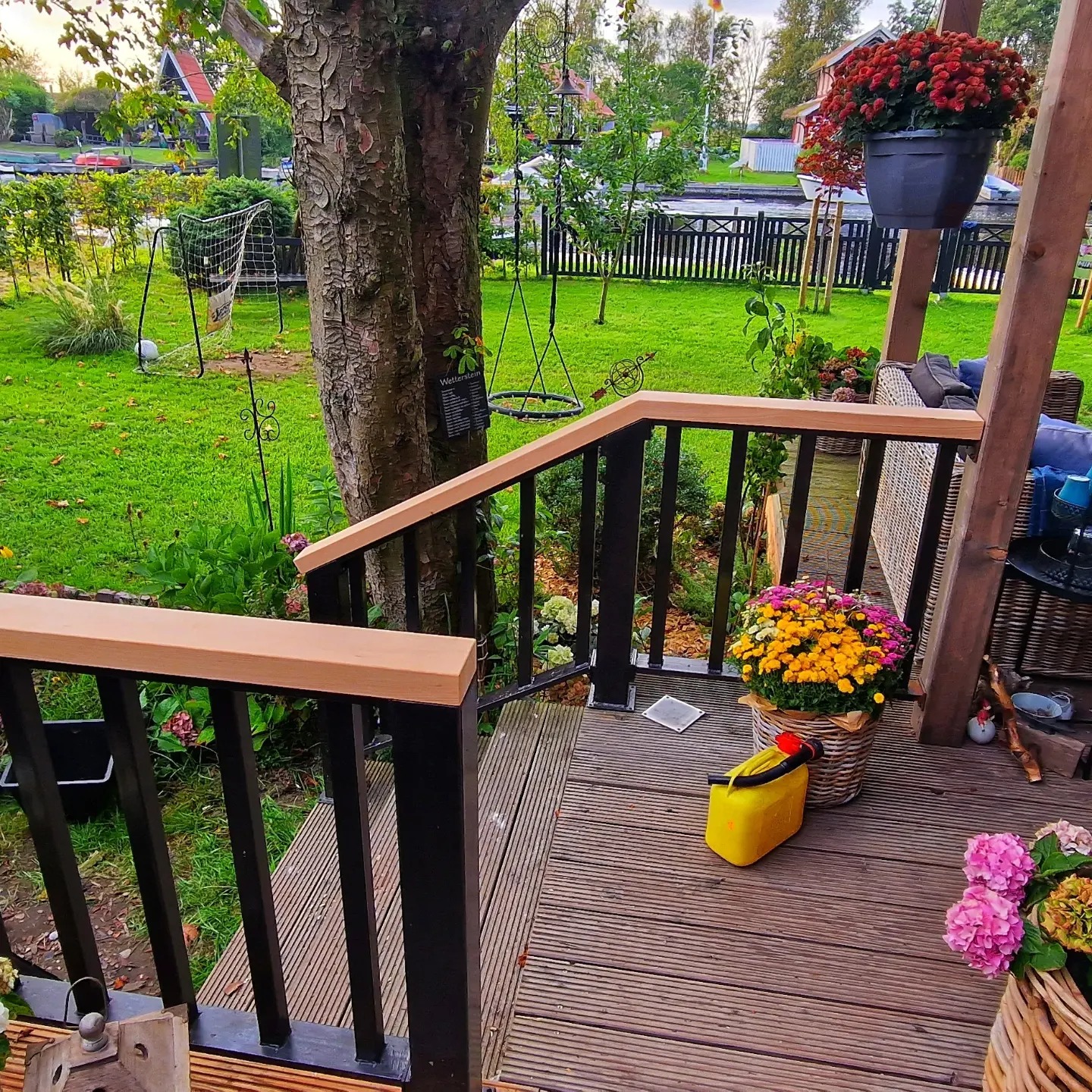 Balustrada z czarnymi słupkami i jasnobrązowymi poręczami na tarasie w Nowym Tomyślu, z widokiem na zielony ogród i drzewo, kwiaty w koszach i donicach dodają koloru.