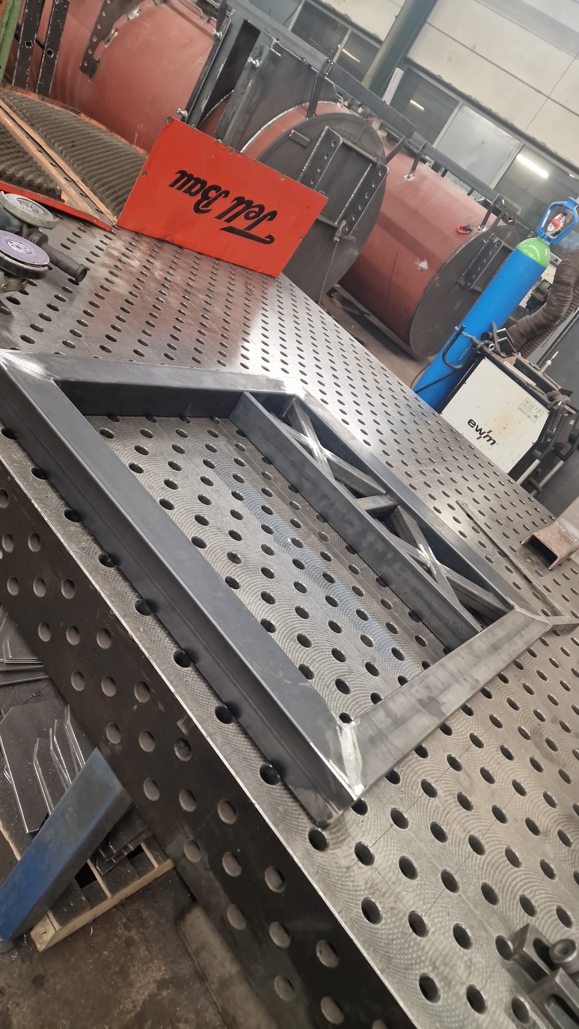 Metalowa rama z poprzeczkami, spawana na perforowanym stole spawalniczym, widoczny sprzęt spawalniczy w tle.