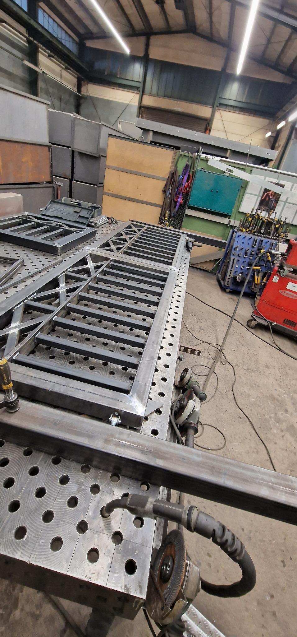 Konstrukcja metalowa, prawdopodobnie fragment balustrady lub bramy, spawana na stole spawalniczym z widocznymi narzędziami szlifierskimi i spawalniczymi w tle hali produkcyjnej.