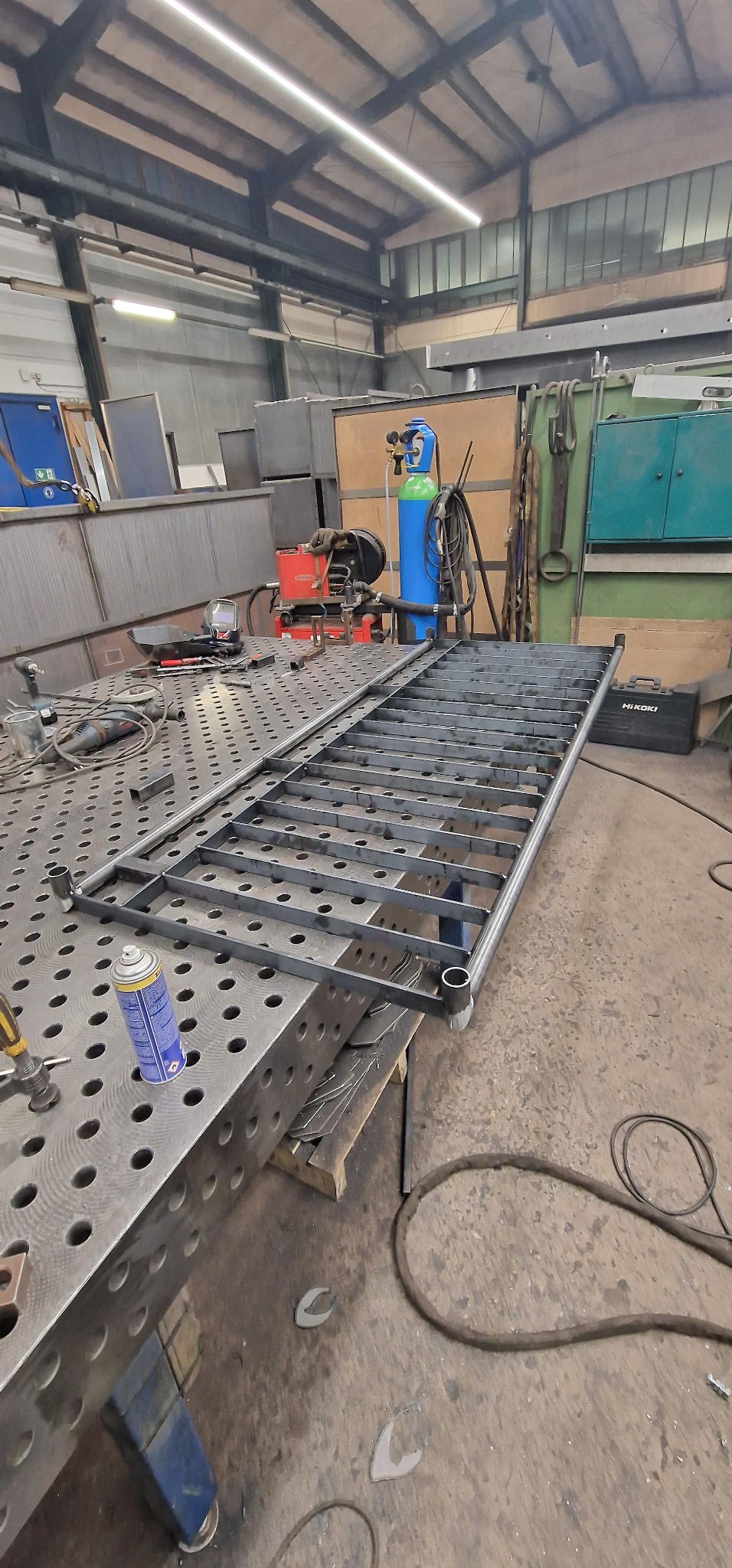 Konstrukcja metalowa balustrady w trakcie spawania na stole spawalniczym z widocznymi narzędziami ślusarskimi i butlą gazową w tle.