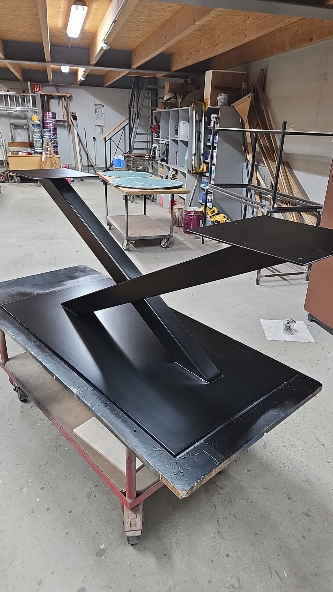 Czarny, metalowy stół z geometryczną podstawą na wózku w warsztacie, widoczne ślady obróbki.