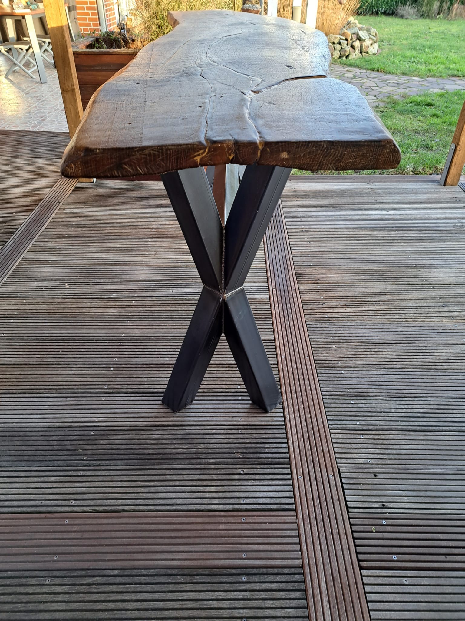 Stół z blatem z litego drewna o nieregularnym kształcie, wsparty na metalowej podstawie w kształcie litery X, ustawiony na drewnianym tarasie.