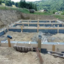 Wykop pod fundamenty domu z widocznym szalunkiem z desek i bloczkami betonowymi, w tle wzgórza zalesione drzewami.