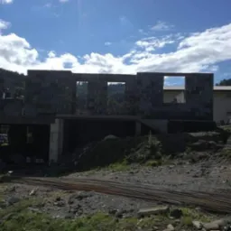 Surowy stan domu w trakcie budowy: ściany z bloczków betonowych z widocznymi otworami okiennymi, zbrojenie na pierwszym planie, górzysty krajobraz w tle pod błękitnym niebem z chmurami.