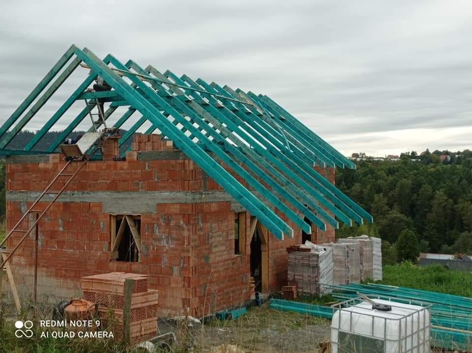 Surowy stan domu w trakcie budowy z czerwonej cegły i z niebieską konstrukcją dachu na tle zalesionego wzgórza pod pochmurnym niebem.