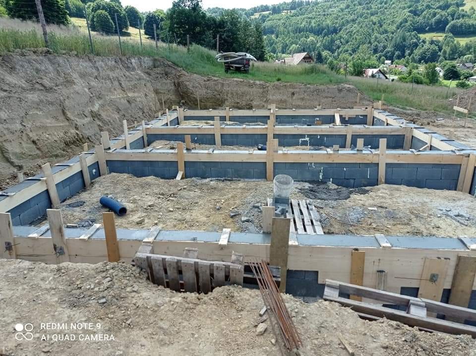 Wykop pod fundamenty domu z widocznym szalunkiem z desek i bloczkami betonowymi, w tle wzgórza zalesione drzewami.