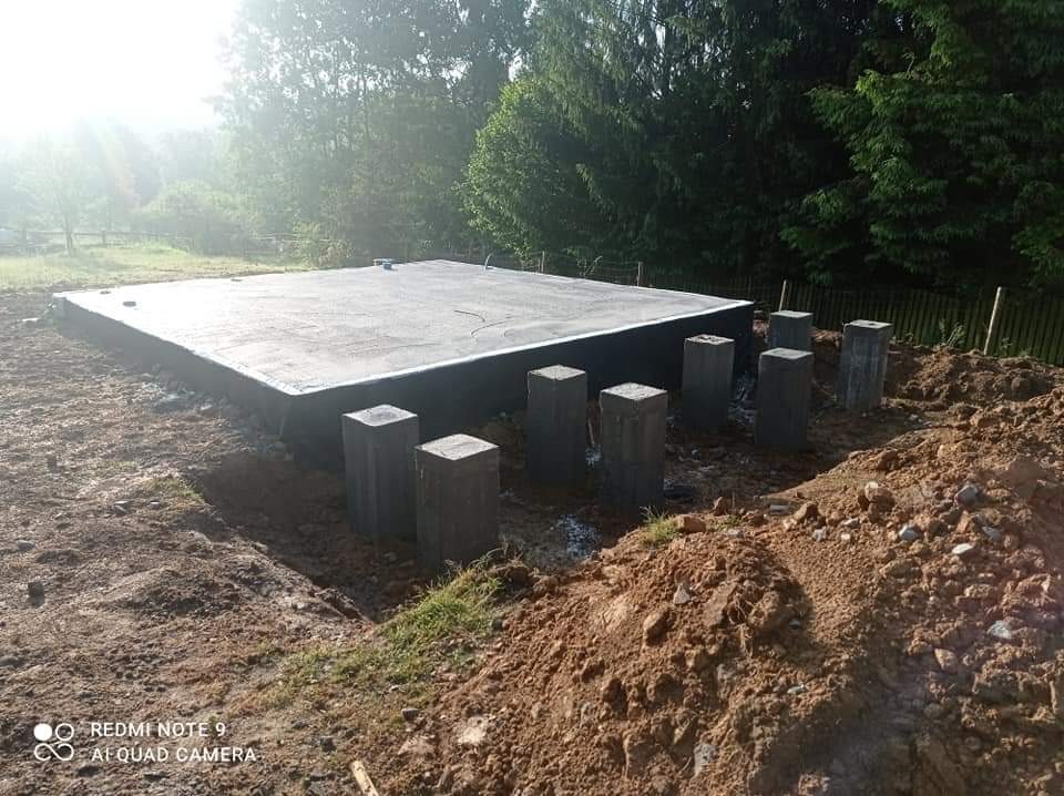 Wylana płyta fundamentowa z izolacją pionową, otoczona betonowymi słupkami i wykopaną ziemią w słoneczny dzień.