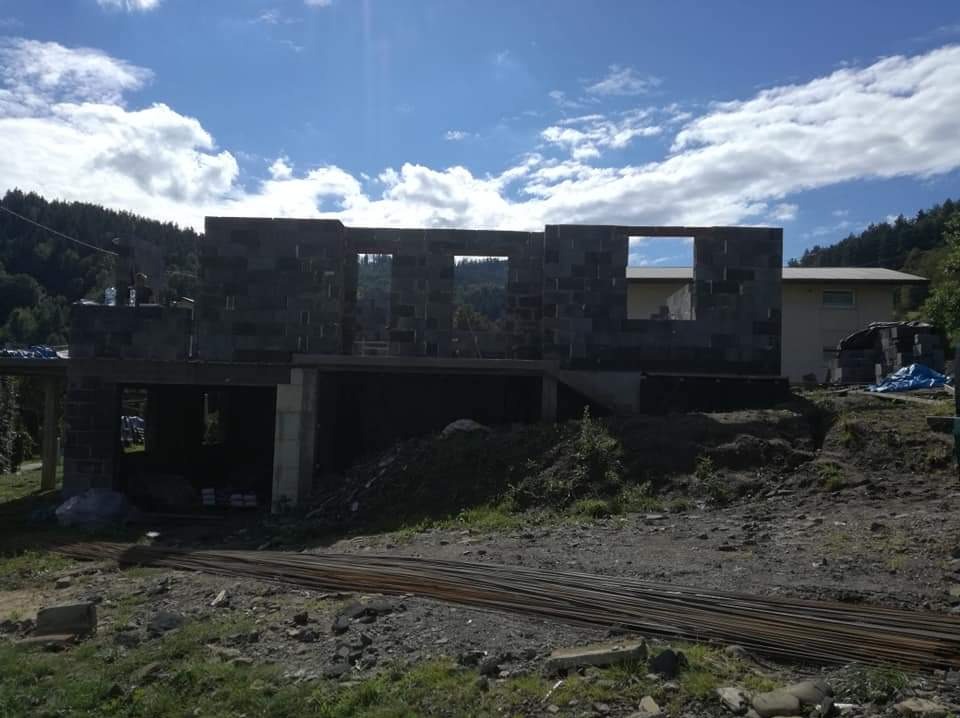 Surowy stan domu w trakcie budowy: ściany z bloczków betonowych z widocznymi otworami okiennymi, zbrojenie na pierwszym planie, górzysty krajobraz w tle pod błękitnym niebem z chmurami.