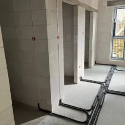 Wnętrze w trakcie prac elektrycznych: widoczne kable w peszlach ułożone na podłodze i wyprowadzone przewody w ścianach z betonu komórkowego. Okno w tle.