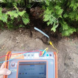 Electro-Safe Adam Jackowski - Miernik rezystancji uziomu w dłoni, wbita sonda w ziemię pod tujami. Ekran miernika pokazuje wynik pomiaru. Przewody pomiarowe w kolorach: czerwony, niebieski, żółty.