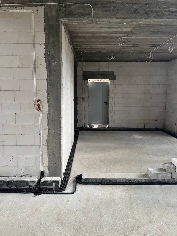 Wnętrze w stanie surowym z widoczną instalacją elektryczną: kable prowadzone po ścianach i pod sufitem, puszki elektryczne. Czarna rura instalacyjna wzdłuż podłogi.