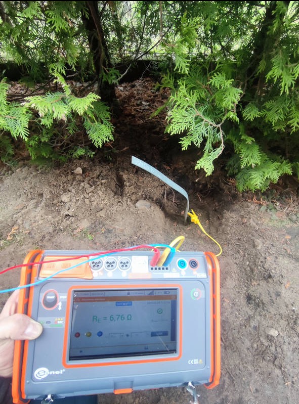 Miernik rezystancji uziomu w dłoni, wbita sonda w ziemię pod tujami. Ekran miernika pokazuje wynik pomiaru. Przewody pomiarowe w kolorach: czerwony, niebieski, żółty.