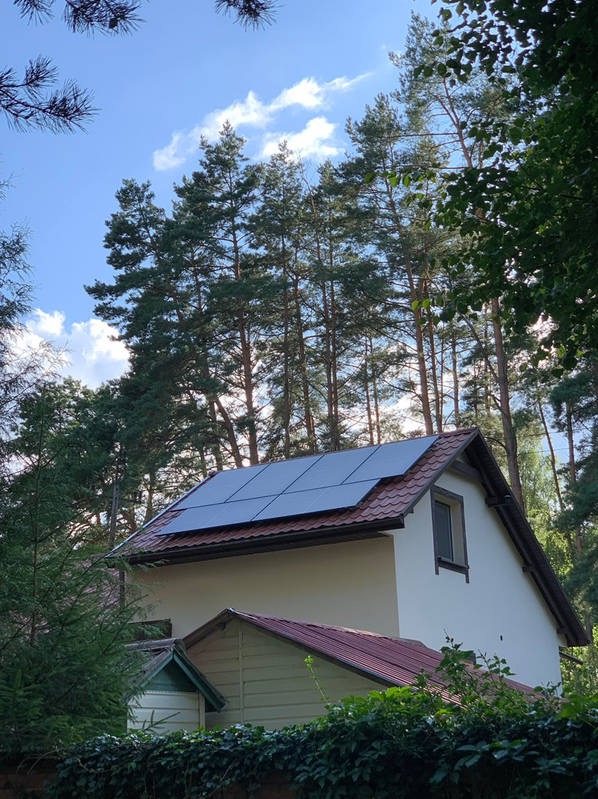 Dom jednorodzinny z panelami fotowoltaicznymi na dachu, otoczony zielenią drzew. Panele wkomponowane w czerwoną dachówkę. Błękitne niebo w tle.