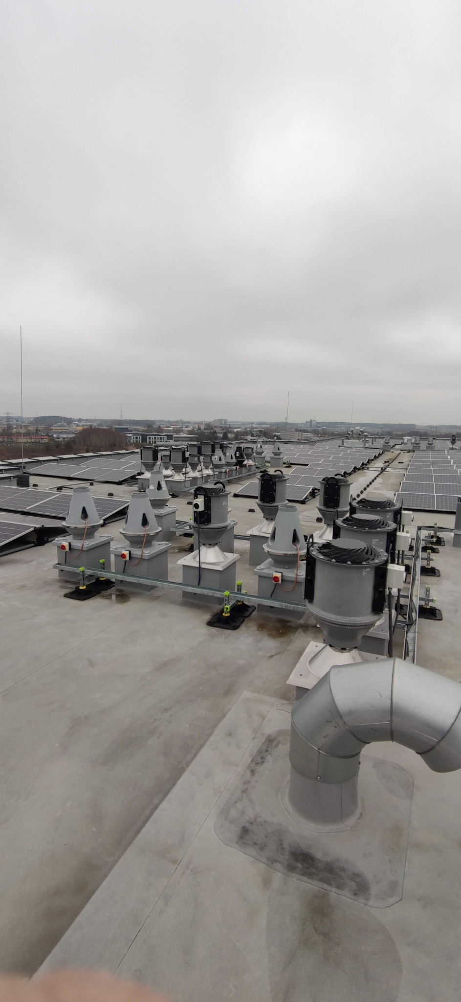 Instalacja fotowoltaiczna i wentylacyjna na dachu budynku przemysłowego w pochmurny dzień. Widoczne panele słoneczne i system wentylacji.