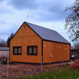 Nowoczesny, drewniany budynek z metalowym dachem, oświetlony zewnętrznymi lampkami, z widoczną skrzynką elektryczną na elewacji i elementami ogrodzenia w tle.