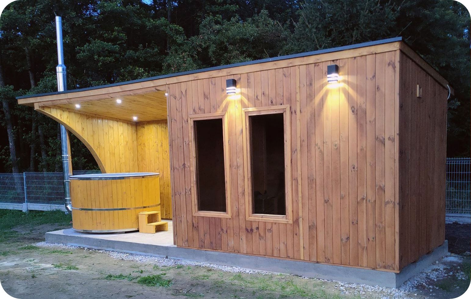 Drewniana konstrukcja z dwoma oknami i dobudowaną wiatą, pod którą znajduje się beczka z wodą i schodkami, oświetlona lampami zewnętrznymi.
