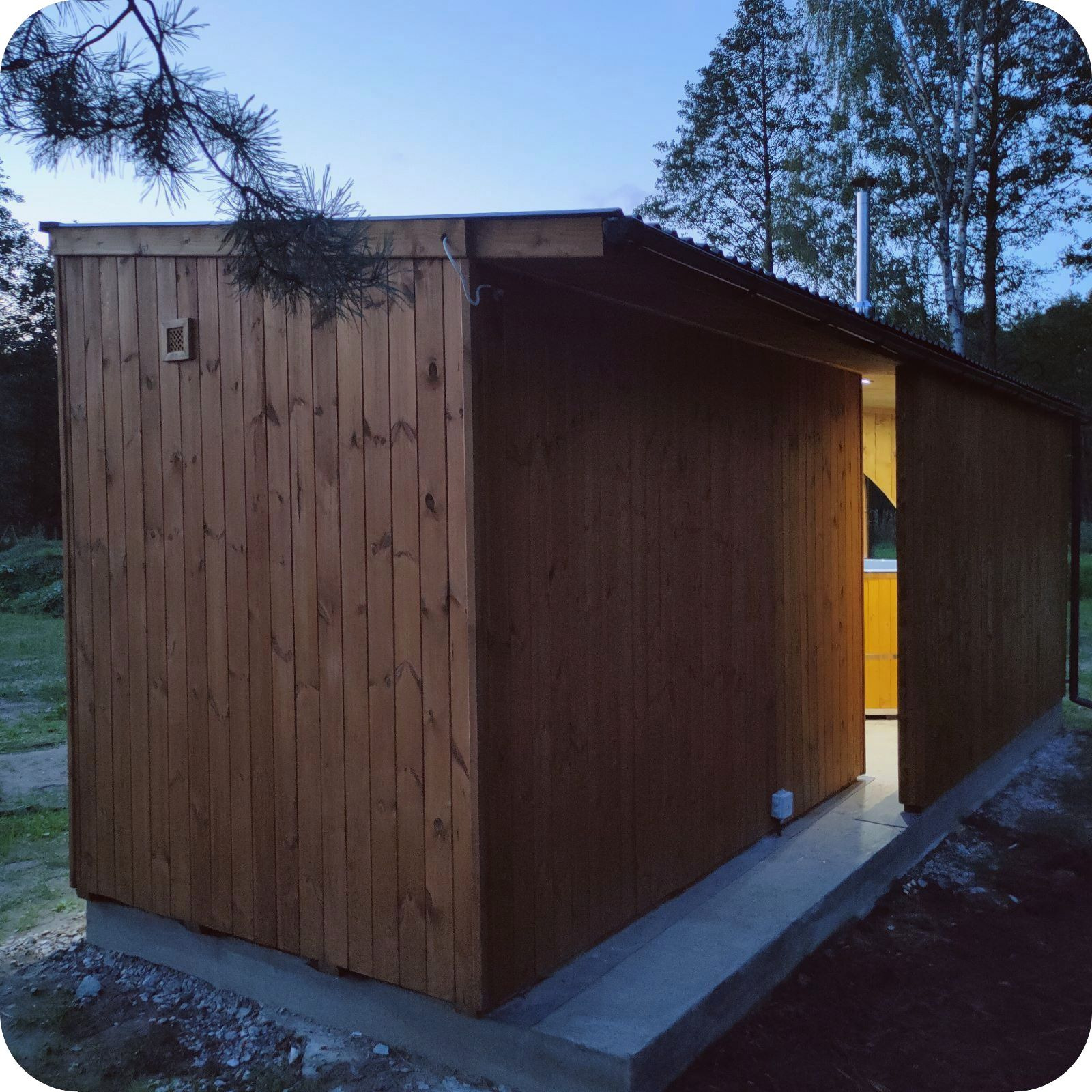 Niewielka, drewniana konstrukcja z kominem, oświetlona od wewnątrz, osadzona na betonowej podstawie, widoczna na tle drzew i nieba o zmierzchu.