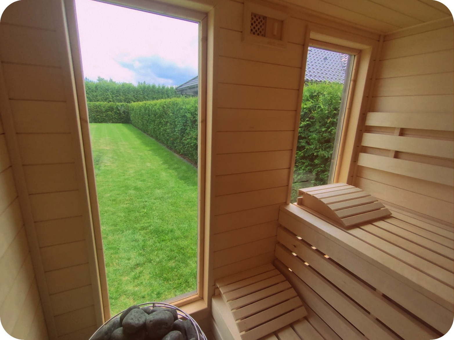 Wnętrze drewnianej sauny z widokiem na zadbany trawnik i żywopłot przez okno, z kamieniami do sauny w metalowym koszu w rogu.