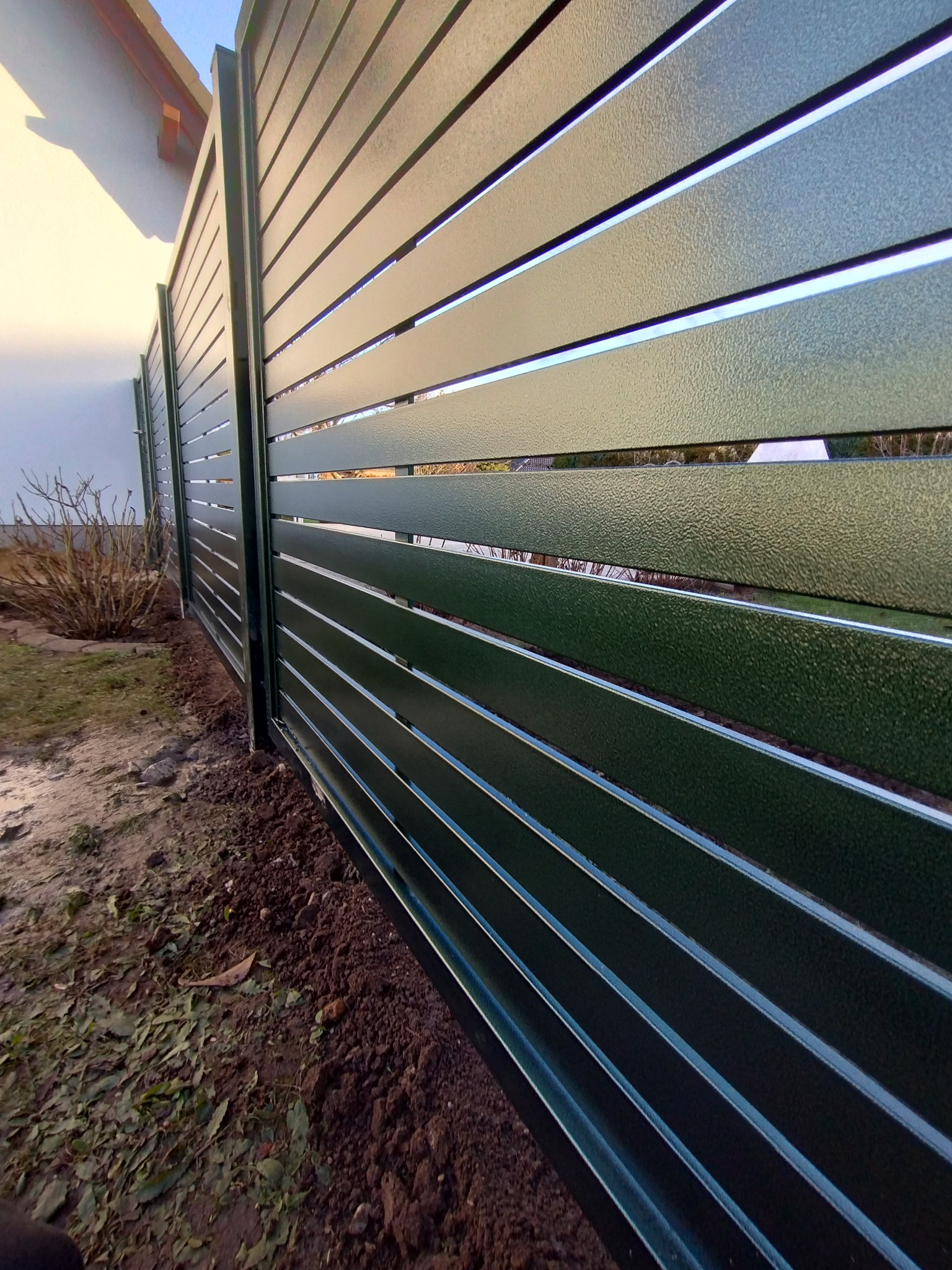 Nowoczesne ogrodzenie panelowe w kolorze ciemnej zieleni, z poziomymi lamelami, widoczne z perspektywy gruntu przy nowo obsypanej ziemi.