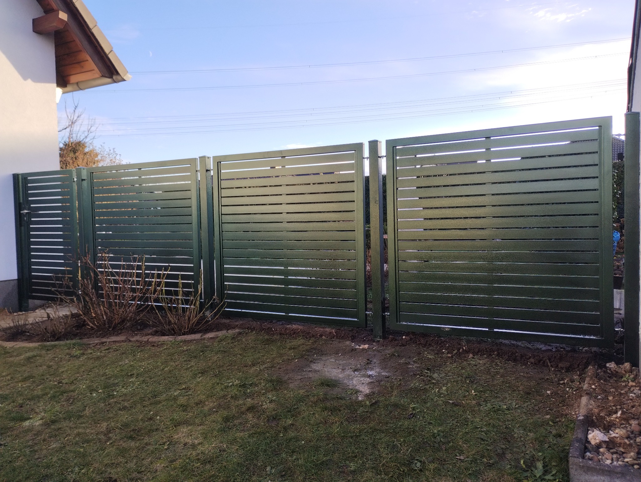 Nowoczesne, poziome ogrodzenie panelowe w kolorze ciemnej zieleni, z szerokimi szczelinami, oddzielające posesję, widoczne krzewy i trawnik, błękitne niebo w tle.