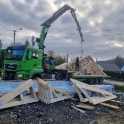 JD&KS SPÓŁKA Z OGRANICZONĄ ODPOWIEDZIALNOŚCIĄ - Montaż dachu z drewnianych wiązarów kratowych za pomocą dźwigu samochodowego MAN. Elementy konstrukcji leżą na ziemi, w tle domy i góry.