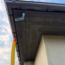 JD&KS SPÓŁKA Z OGRANICZONĄ ODPOWIEDZIALNOŚCIĄ - Zamontowany, ciemny system rynnowy z deską okapową. Widoczny fragment ściany budynku i żółta rura spustowa. Błękitne niebo w tle.