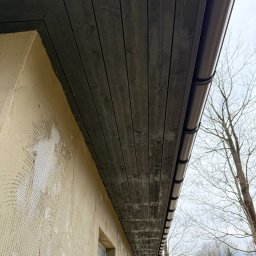 JD&KS SPÓŁKA Z OGRANICZONĄ ODPOWIEDZIALNOŚCIĄ - Wykończenie okapu dachu z drewnianą podbitką i rynnami w kolorze brązowym, ściana budynku z siatką pod tynk. Widok z dołu, z fragmentem krajobrazu w tle.
