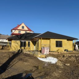 JD&KS SPÓŁKA Z OGRANICZONĄ ODPOWIEDZIALNOŚCIĄ - Budowa domu w stanie surowym otwartym: żółte ściany z izolacją, czarny dach, okna. Na pierwszym planie ziemia i materiały budowlane.