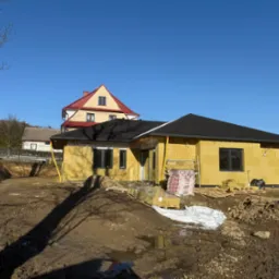 Budowa domu w trakcie: ściany z izolacją w żółtym kolorze, czarny dach, teren wokół w stanie surowym. Widoczny materiał budowlany i rusztowanie. Słoneczny dzień.