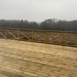 Drewniany taras z balustradą w stylu rustykalnym, widok na pole i las w mglisty dzień. Deski tarasowe z widocznym usłojeniem. Balustrada z krzyżowymi elementami.