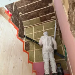 Pracownik w kombinezonie ochronnym poziomuje ścianę z izolacją w surowym wnętrzu domu w trakcie budowy. Widoczne drewniane elementy konstrukcyjne.