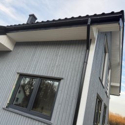 JD&KS SPÓŁKA Z OGRANICZONĄ ODPOWIEDZIALNOŚCIĄ - Szary dom z drewnianą elewacją i czarnymi oknami, rynnami. Widok z dołu na fragment budynku z widocznym fragmentem dachu i niebem w tle.