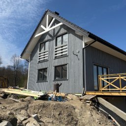 JD&KS SPÓŁKA Z OGRANICZONĄ ODPOWIEDZIALNOŚCIĄ - Szary dom z drewnianą elewacją, balkonem i białymi detalami architektonicznymi. Na pierwszym planie ziemia, materiały budowlane i fragment wykopu pod fundament.