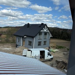 JD&KS SPÓŁKA Z OGRANICZONĄ ODPOWIEDZIALNOŚCIĄ - Szary dom z czarnym dachem w trakcie budowy, widok z góry, na pierwszym planie fragment czerwonego dachu i biały bus z otwartymi drzwiami.