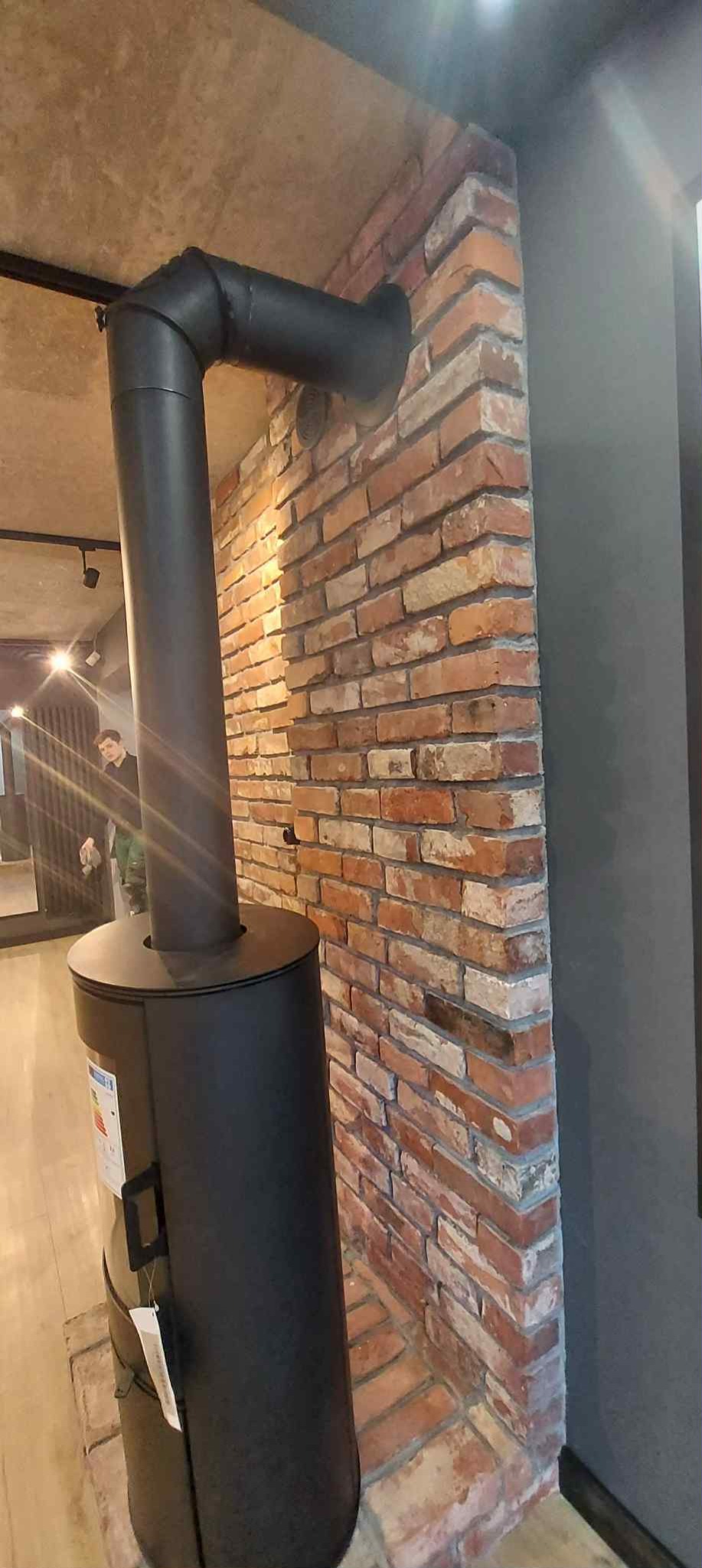 Ceglana ściana z kominem w nowoczesnym wnętrzu. Widoczny fragment sufitu i podłogi. W tle osoba w zielonej bluzie.