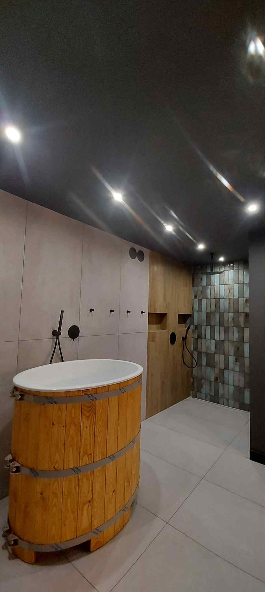 Nowoczesna łazienka z drewnianą wanną beczką, szarą podłogą i oświetleniem punktowym. Ściany wykończone płytkami w różnych odcieniach i fakturach.