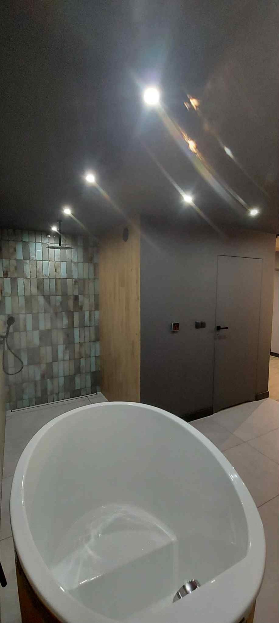 Nowoczesna łazienka z wolnostojącą wanną, mozaiką na ścianie prysznica i punktowym oświetleniem sufitowym. Minimalistyczny design, szare ściany i drewniane akcenty.